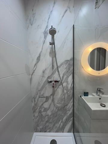 Nowoczesna kabina prysznicowa z marmurowymi płytkami, szklaną ścianką i okrągłym lustrem z podświetleniem LED. Minimalistyczny design, czyste linie, jasne kolory.