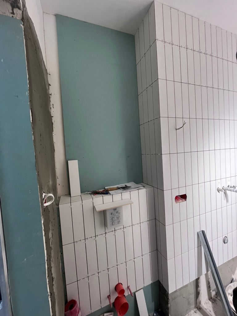 Białe, pionowe płytki w trakcie układania w łazience, widoczne narzędzia i elementy instalacji. Ściana z płytą karton-gips w tle. Etap prac wykończeniowych.