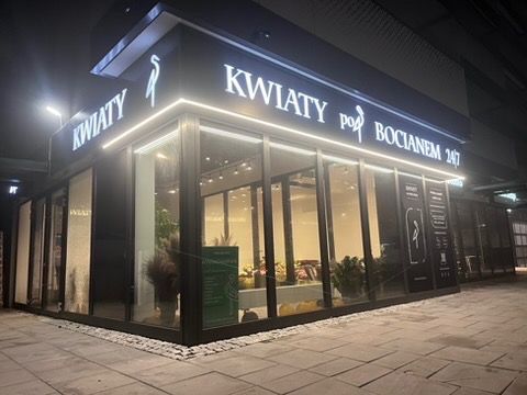 Nowoczesny pawilon kwiaciarni z dużymi przeszkleniami i podświetlanym szyldem 'KWIATY pod BOCIANEM 24/7' nocą. Widoczne kwiaty i dekoracje wewnątrz.