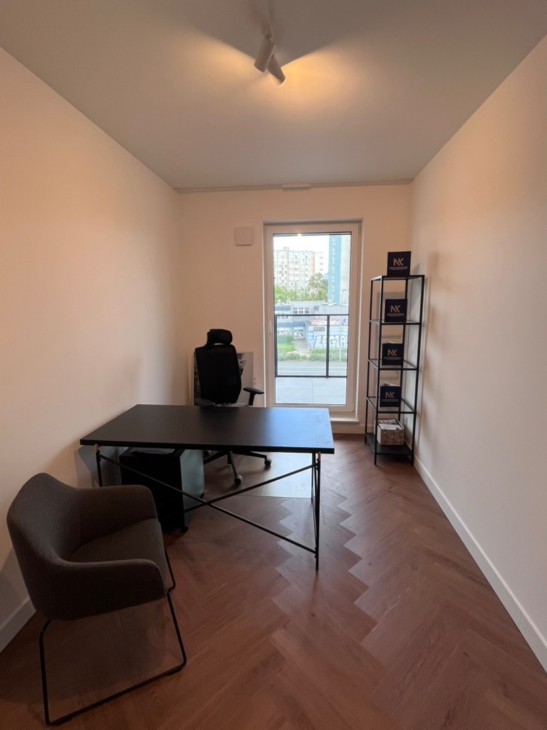 Nowoczesne biuro z parkietem w jodełkę, czarnym biurkiem i krzesłem. Minimalistyczny regał z logo firmy przy oknie. Jasne ściany i sufit z oświetleniem punktowym.