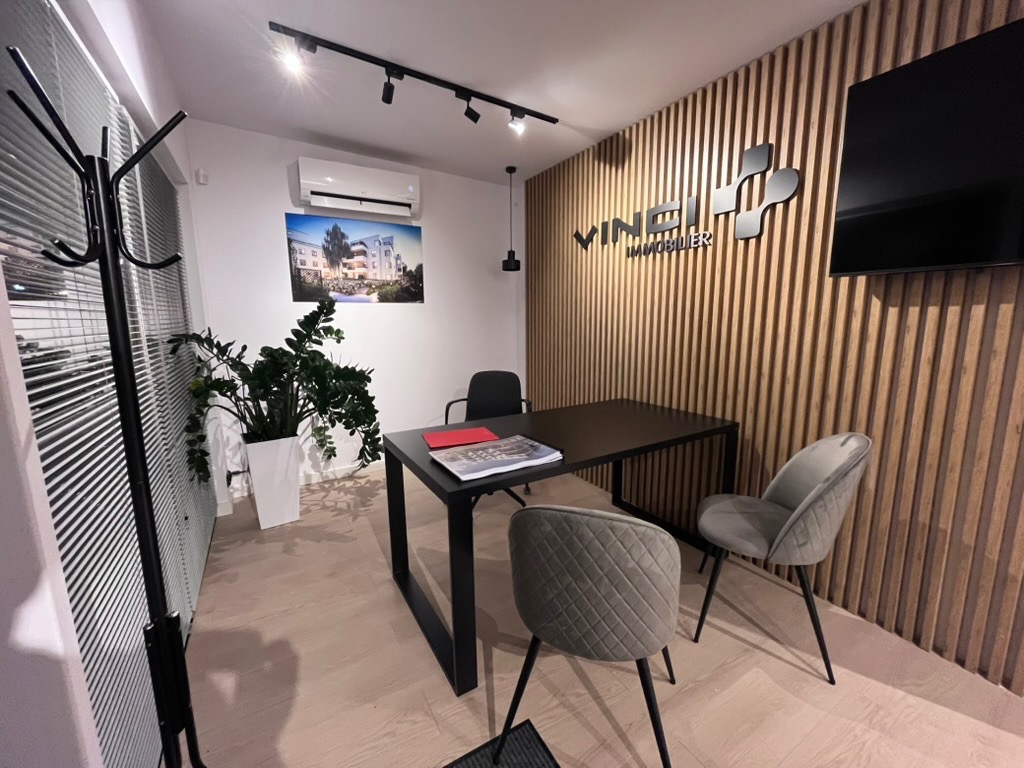 Nowoczesne biuro z drewnianą ścianą, czarnym stołem i szarymi krzesłami. Na ścianie logo firmy i telewizor. Minimalistyczny design z roślinami i oświetleniem punktowym.