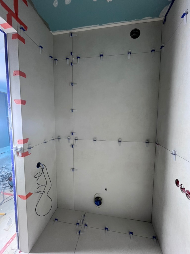 Ściany w trakcie układania płytek w łazience, z użyciem systemu poziomowania. Widoczne przewody elektryczne i otwory na instalacje. Szare płytki, niebieski sufit.