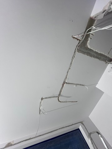 Biały sufit z wystającymi przewodami elektrycznymi i fragmentem uszkodzonego narożnika, widoczne ślady po instalacji oświetlenia.