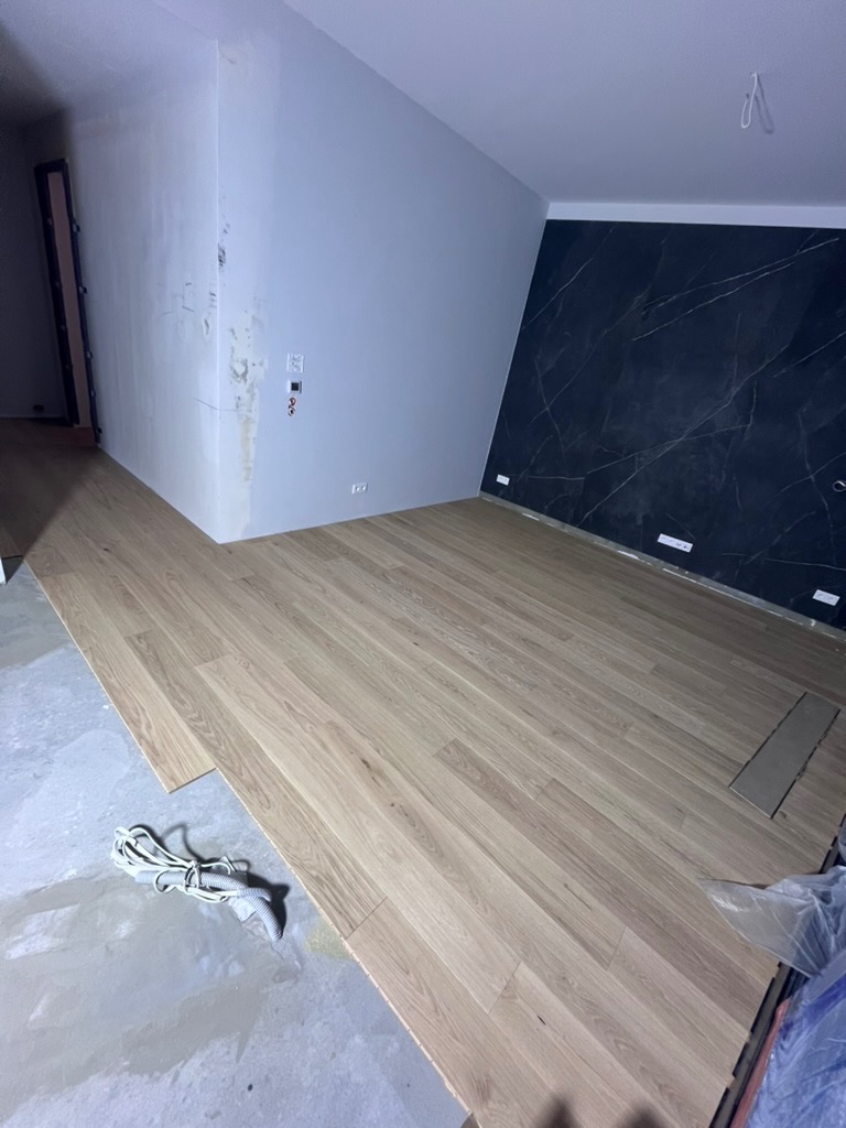 Układanie jasnej drewnianej podłogi w pokoju z szarą ścianą i czarną marmurową ścianą akcentującą. Widoczne narzędzia i materiały budowlane.