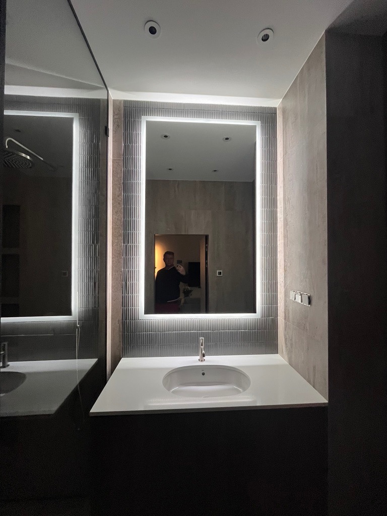 Nowoczesna łazienka z podświetlanym lustrem i umywalką nablatową. Szare płytki i ciemne meble tworzą elegancki kontrast. Widoczne detale wykończenia.