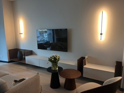 Nowoczesny salon z białymi meblami, telewizorem i minimalistycznymi lampami ściennymi. Bukiet białych tulipanów na stoliku kawowym. Jasna podłoga i neutralne kolory ścian.