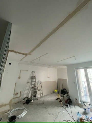Wnętrze w trakcie prac wykończeniowych: szpachlowanie sufitu, widoczne profile, drabiny, narzędzia i materiały budowlane. Prace w jasnym pomieszczeniu z oknem.