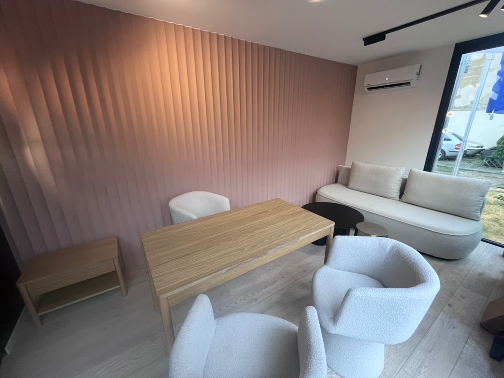 Nowoczesne wnętrze z teksturowaną, różową ścianą, drewnianym stołem i białymi fotelami. Widoczny fragment sofy i okna z widokiem na zewnątrz. Minimalistyczny design.