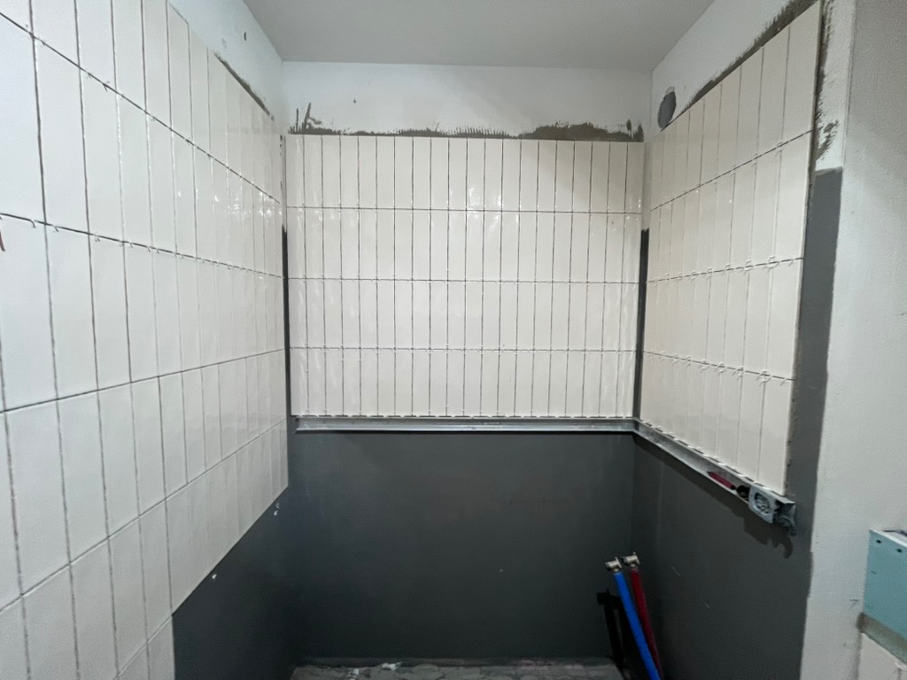 Wnętrze łazienki w trakcie wykańczania: białe, błyszczące płytki ułożone na ścianach do połowy wysokości, szara farba poniżej, widoczne rurki instalacyjne.