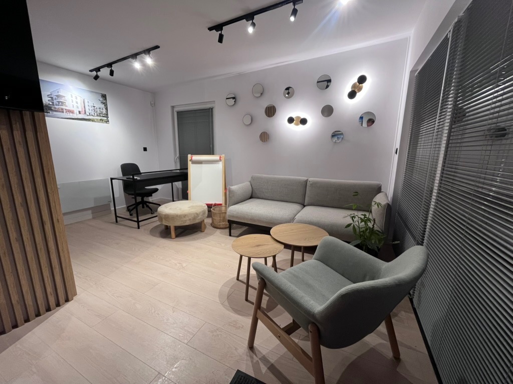 Jasne, nowocześnie urządzone wnętrze biura z drewnianą podłogą, szarą sofą i designerskimi lampami na ścianie. Widoczne biurko i krzesło w tle.