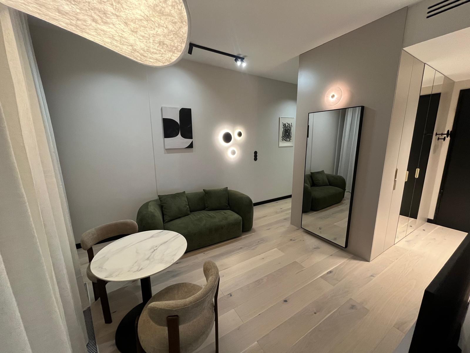 Nowoczesny salon z zieloną sofą, okrągłym stołem z marmurowym blatem i dużym lustrem, odbijającym wnętrze. Jasne drewniane podłogi i designerskie oświetlenie.