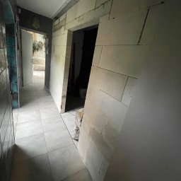 MK REMONT Mateusz Kubisz - Korytarz w trakcie prac wykończeniowych: ułożona szara podłoga z płytek, obudowa drzwi z białych bloków z numerami, widoczny worek z zaprawą.