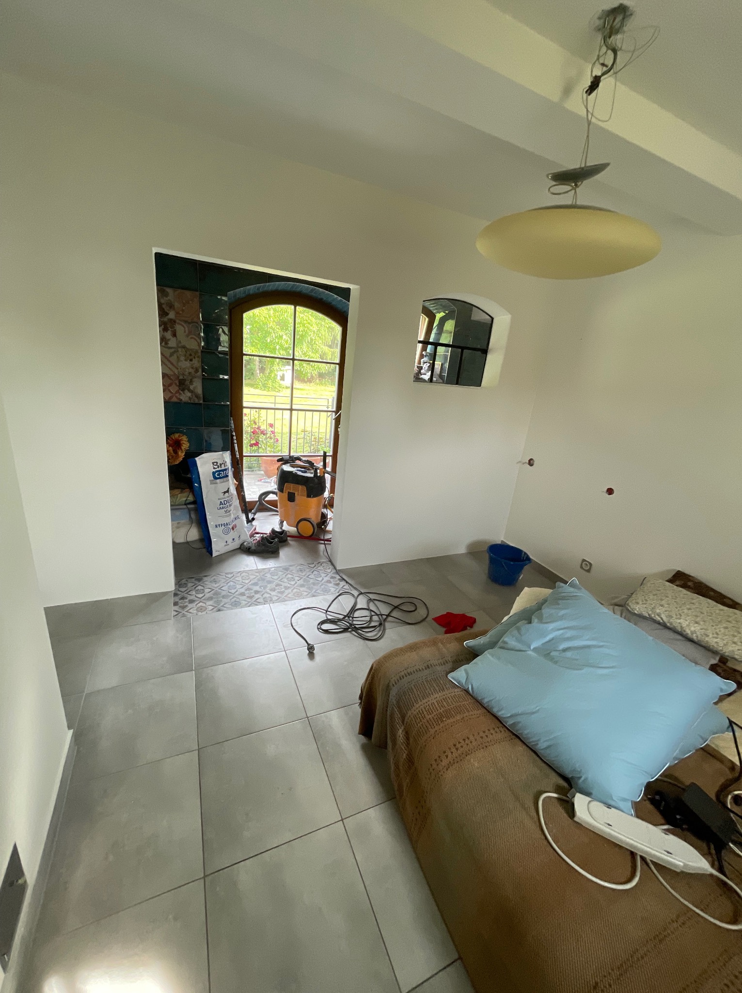 Szare płytki podłogowe w pokoju z widokiem na ogród przez drzwi. W tle torba z klejem i odkurzacz. Częściowo widoczny tapczan z poduszką.