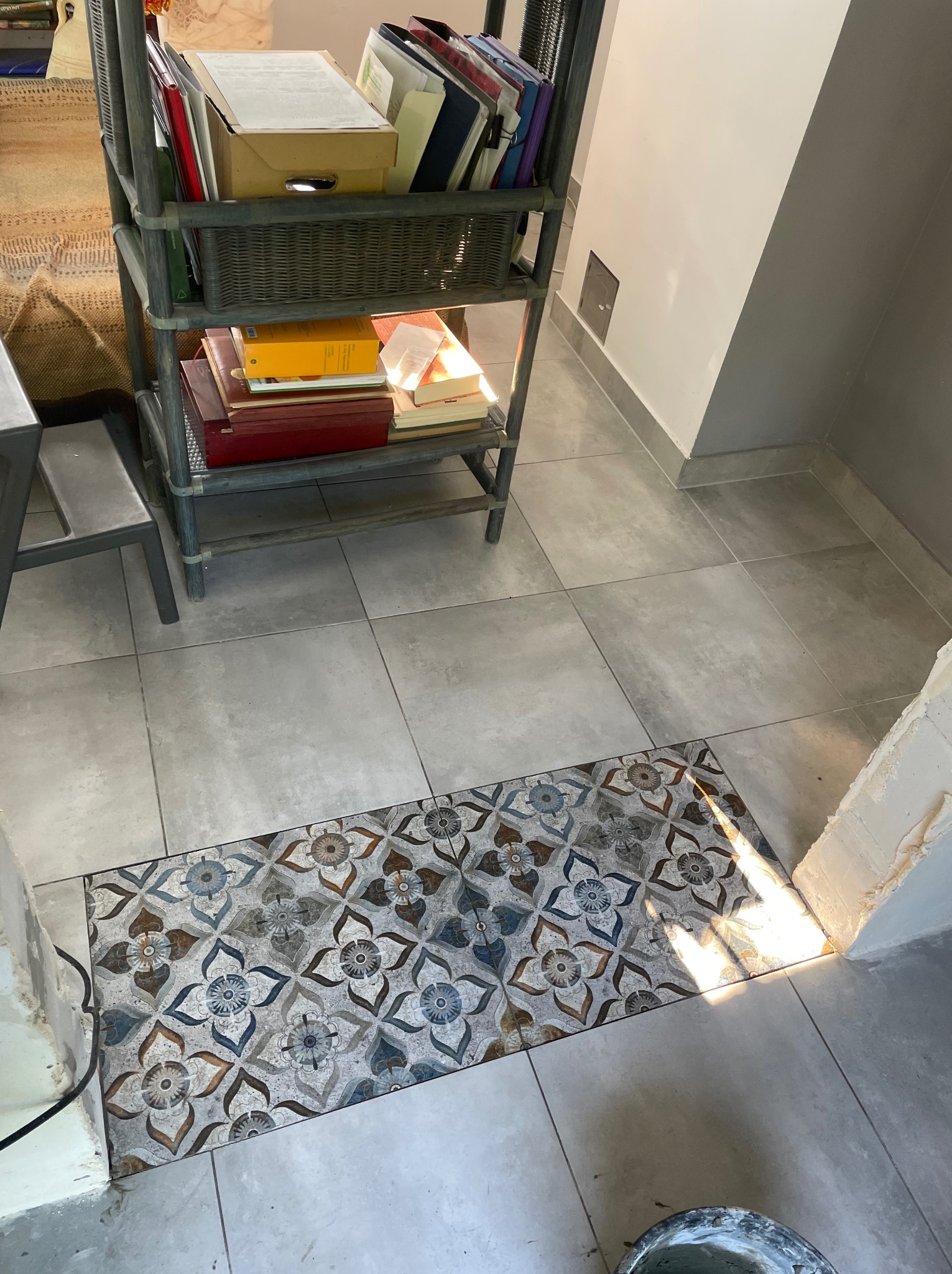 Ułożona, wzorzysta płytka podłogowa w odcieniach szarości, błękitu i brązu, kontrastująca z jasnymi, gładkimi płytkami. Fragment regału z książkami w tle.