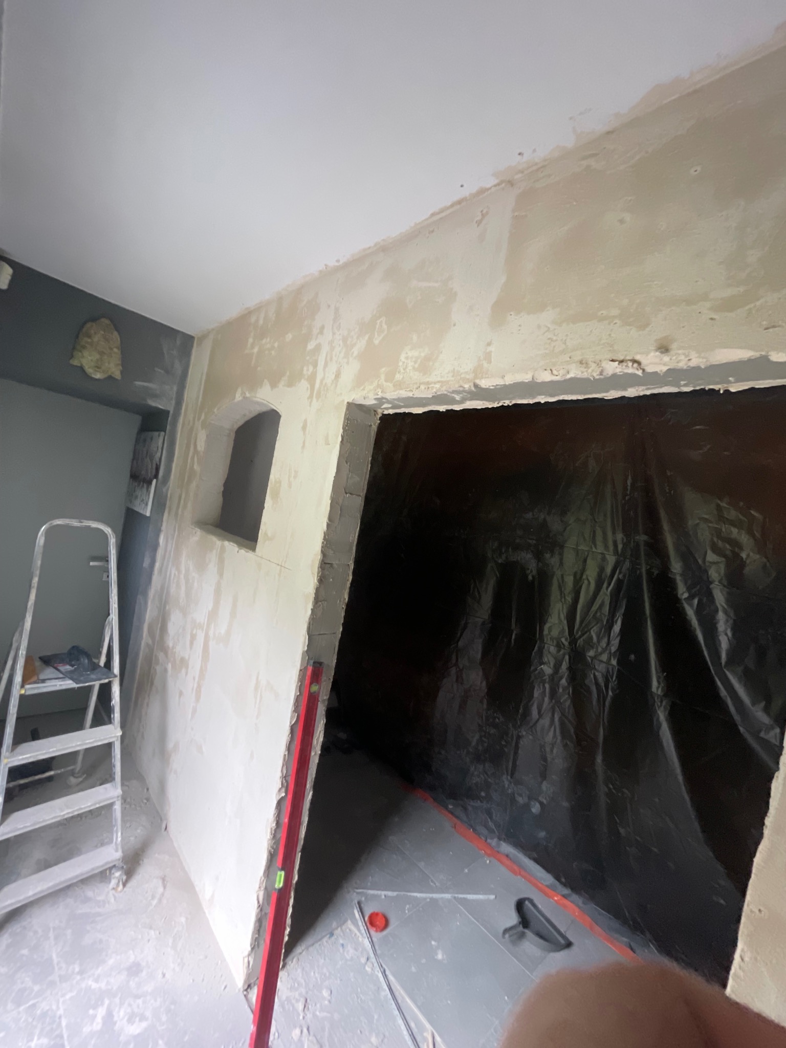 Wnętrze w trakcie remontu: ściany z nałożoną gładzią, drabina, poziomica i folia ochronna. Widoczne przygotowania do dalszych prac wykończeniowych.