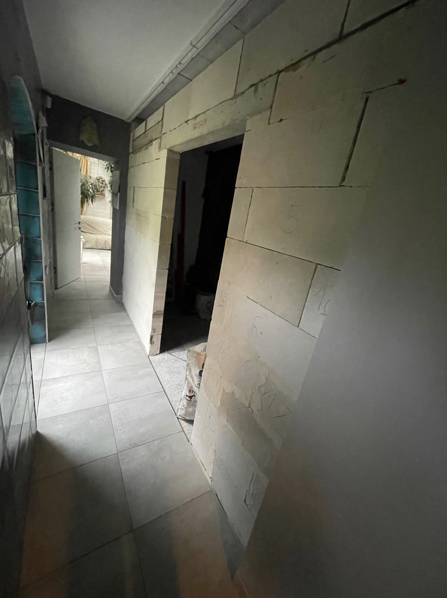 Korytarz w trakcie prac wykończeniowych: ułożona szara podłoga z płytek, obudowa drzwi z białych bloków z numerami, widoczny worek z zaprawą.