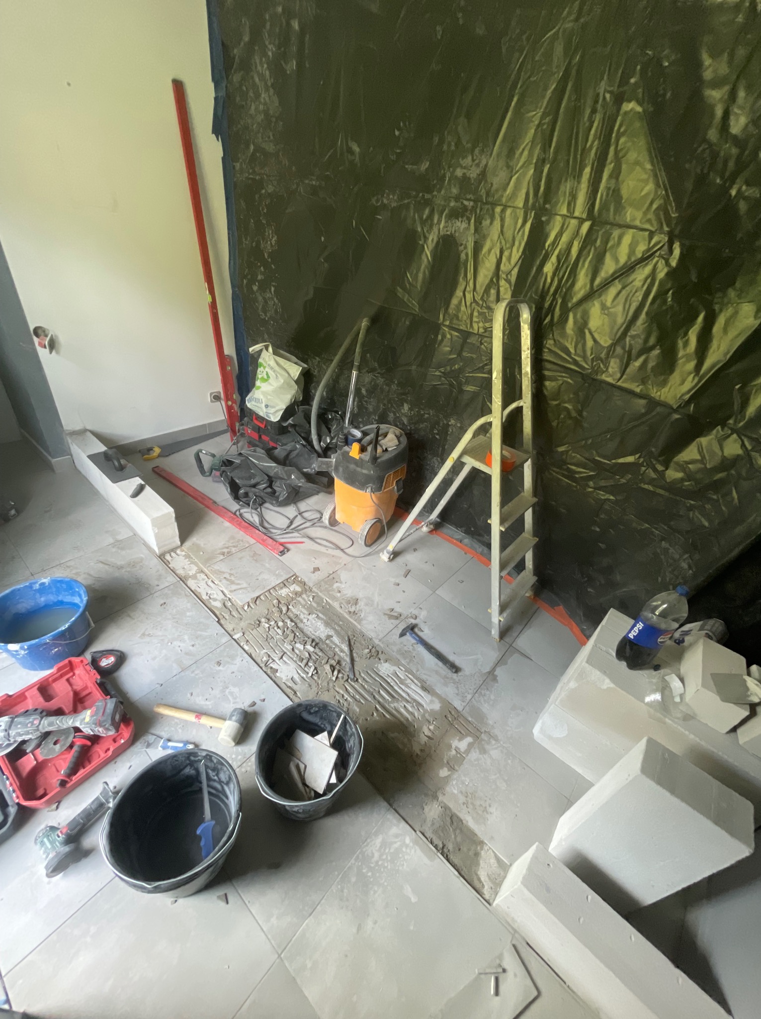Demontaż płytek podłogowych w trakcie prac. Widoczne narzędzia, drabina, wiadro i gruz. Podłoga częściowo odkryta, ściana zabezpieczona folią.