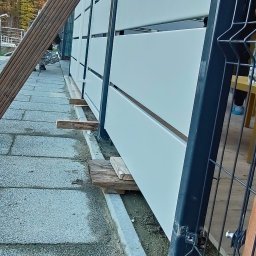 Metalnova - Nowoczesne, białe panele ogrodzeniowe z czarną ramą, w trakcie montażu. Widoczne drewniane podpory i fragment posesji w tle. Ujęcie z dołu, podkreślające perspektywę.