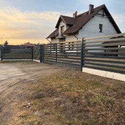 Metalnova - Nowoczesna, pozioma brama przesuwna i ogrodzenie z metalowych profili w kolorze grafitowym, na tle domu jednorodzinnego i zachodzącego słońca.