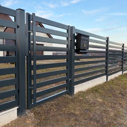 Metalnova - Nowoczesne, poziome, metalowe ogrodzenie w kolorze grafitowym z czarną skrzynką na listy. W tle dom jednorodzinny i błękitne niebo.