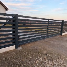 Metalnova - Nowoczesna, pozioma brama wjazdowa w kolorze grafitowym, z widocznymi poziomymi przęsłami. Brama zamontowana na betonowej podmurówce, tło stanowi błękitne niebo.