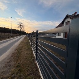 Metalnova - Nowoczesne, poziome ogrodzenie metalowe w kolorze grafitowym, z betonową podmurówką, oddzielające posesję od drogi. W tle dom jednorodzinny i zachodzące słońce.
