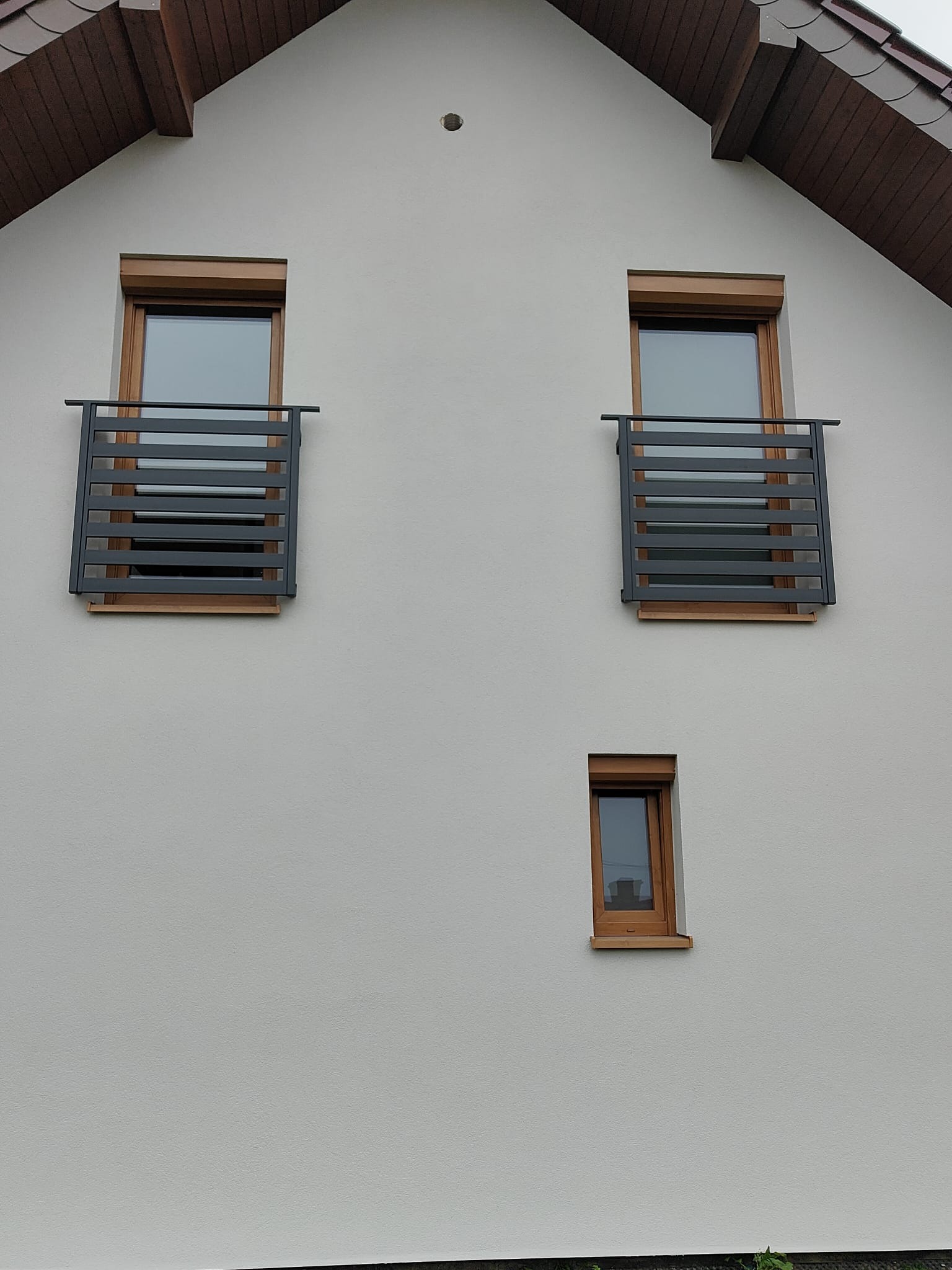 Elewacja domu z dwoma oknami zabezpieczonymi nowoczesnymi, poziomymi balustradami w kolorze grafitowym. Małe okno poniżej. Dach z brązową podbitką.