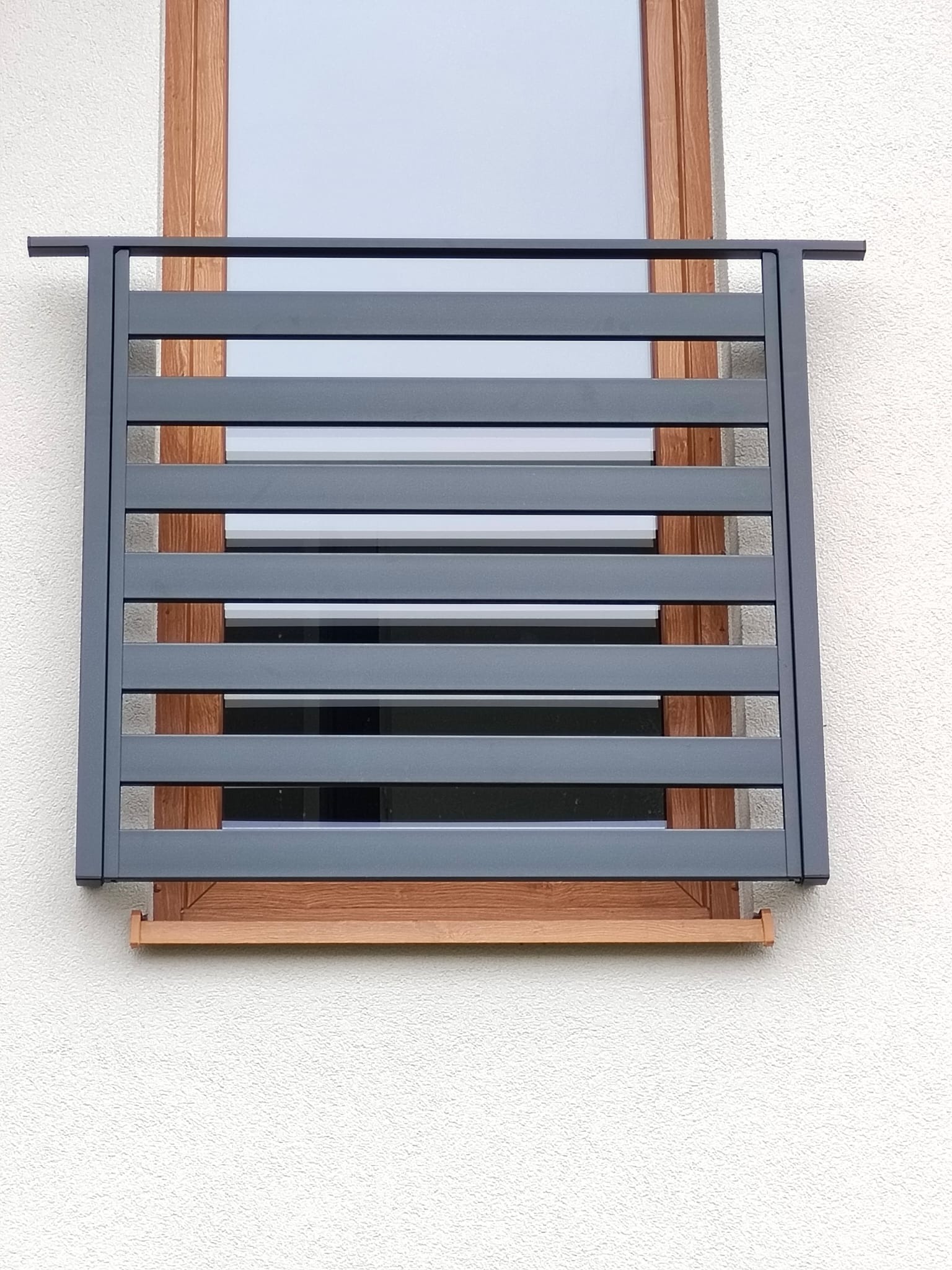 Szara, pozioma osłona okienna z metalu, zamontowana na drewnianej ramie okna na tle białej, teksturowanej ściany zewnętrznej budynku. Minimalistyczny design.