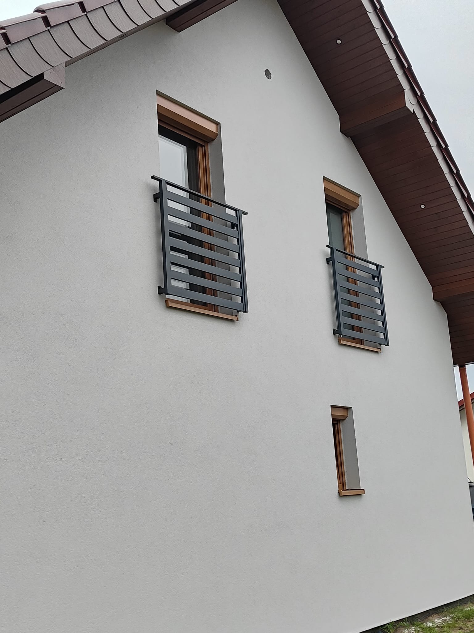 Elewacja domu z oknami zabezpieczonymi nowoczesnymi, poziomymi, metalowymi balustradami w kolorze grafitowym. Widok z dołu, na tle jasnego nieba.