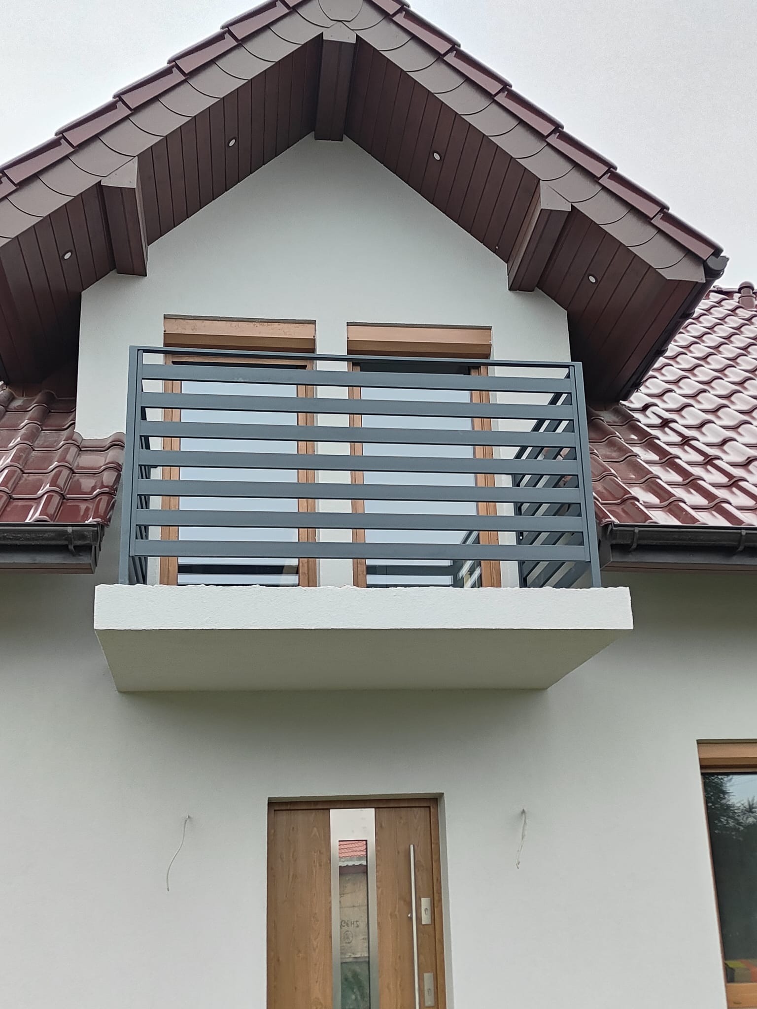 Szara, pozioma balustrada balkonowa z metalu na tle białej elewacji domu z brązowym dachem. Widok z dołu, podkreślający nowoczesny design i solidne wykonanie.
