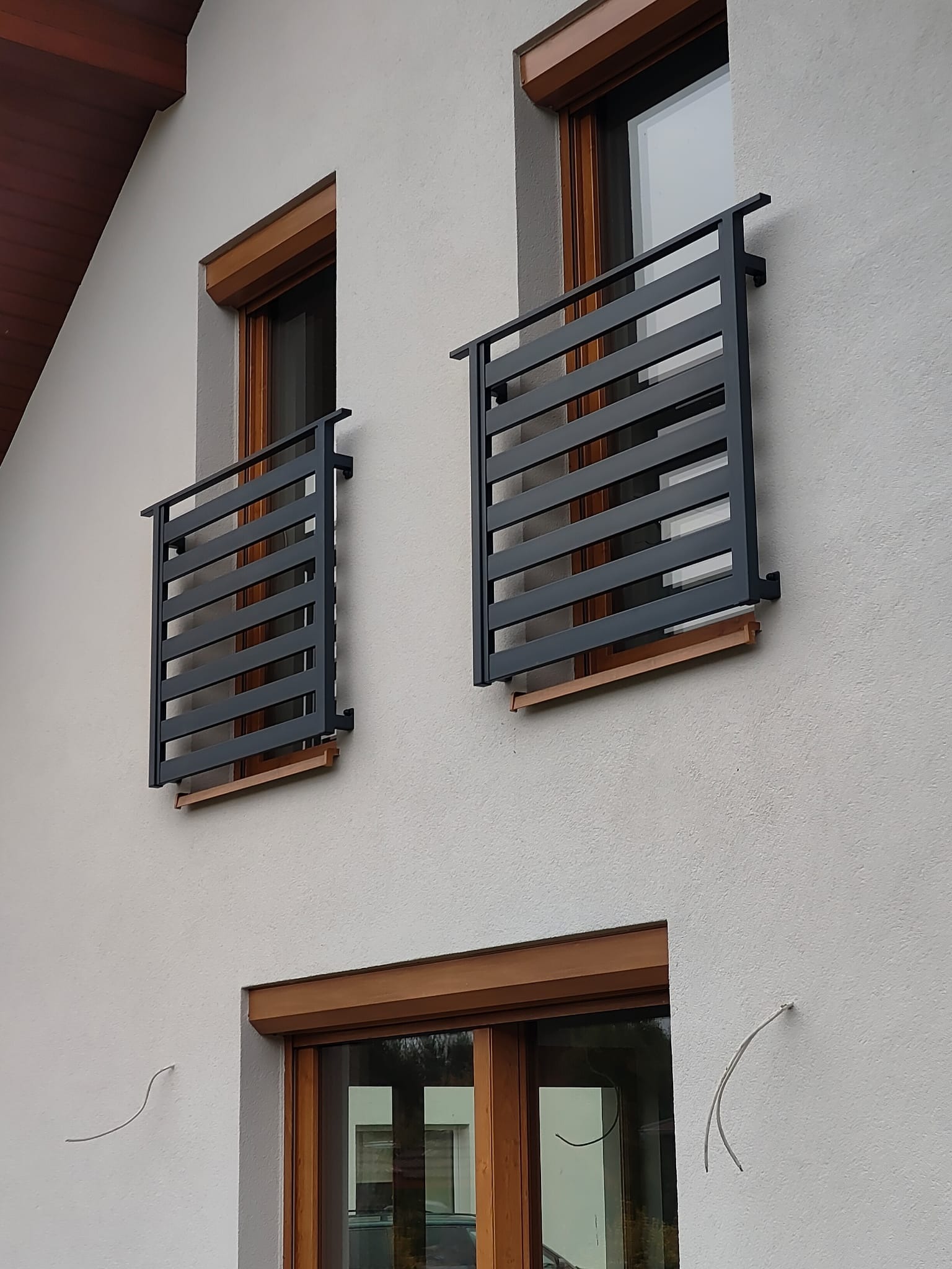 Dwa metalowe, poziome zabezpieczenia okienne w kolorze grafitowym, zamontowane na elewacji budynku z oknami z drewnianą stolarką. Widok z dołu.