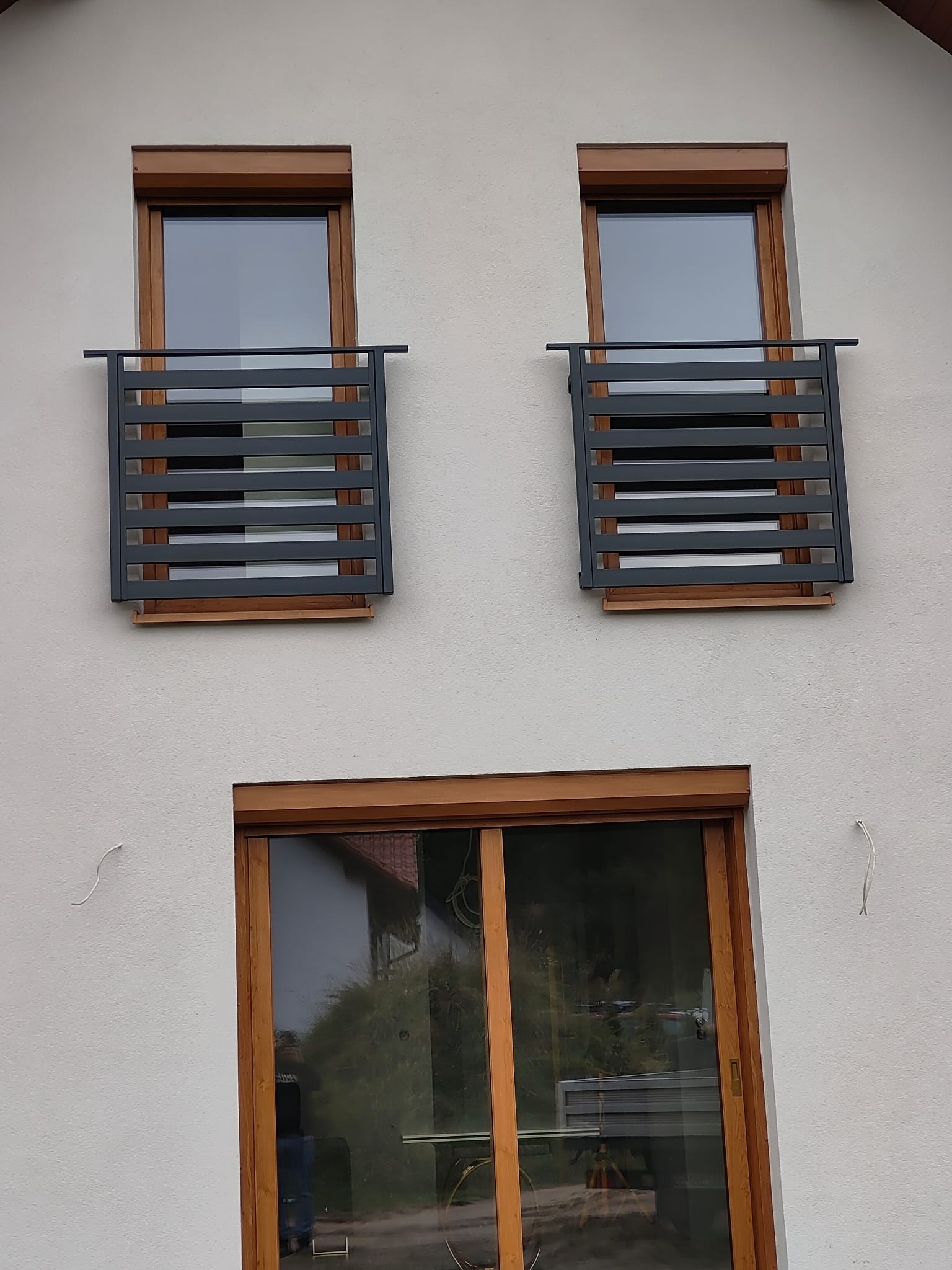 Dwa okna z zamontowanymi, nowoczesnymi, metalowymi balustradami w kolorze grafitowym, z poziomymi szczebelkami. Na dole duże okno przesuwne z drewnianą ramą.