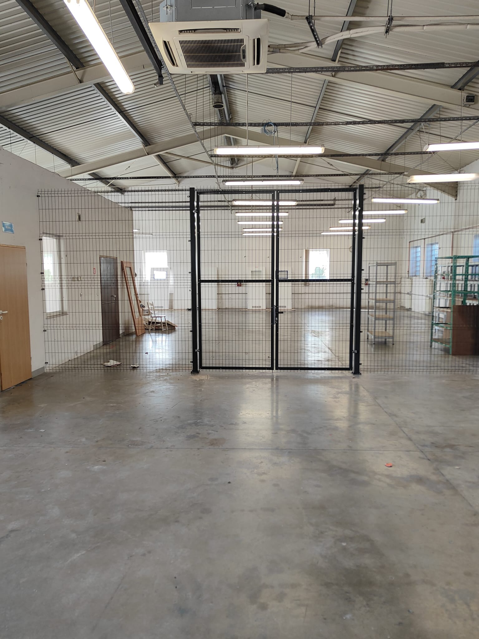 Wnętrze hali z zainstalowanym systemem siatkowych przegród i bram w kolorze czarnym. Widoczne oświetlenie sufitowe i betonowa posadzka. Przegrody dzielą przestrzeń na sekcje.