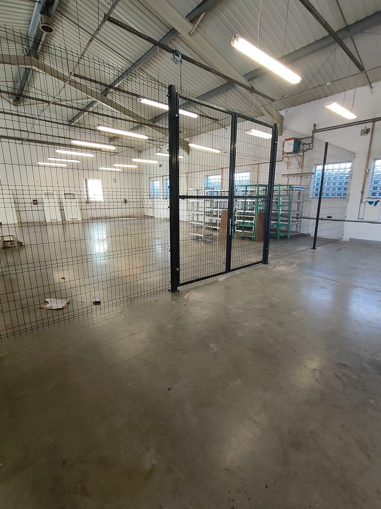 Wnętrze hali z siatkową konstrukcją oddzielającą przestrzeń, widoczne regały i okna z luksferami. Oświetlenie jarzeniowe podkreśla surowy charakter wnętrza.
