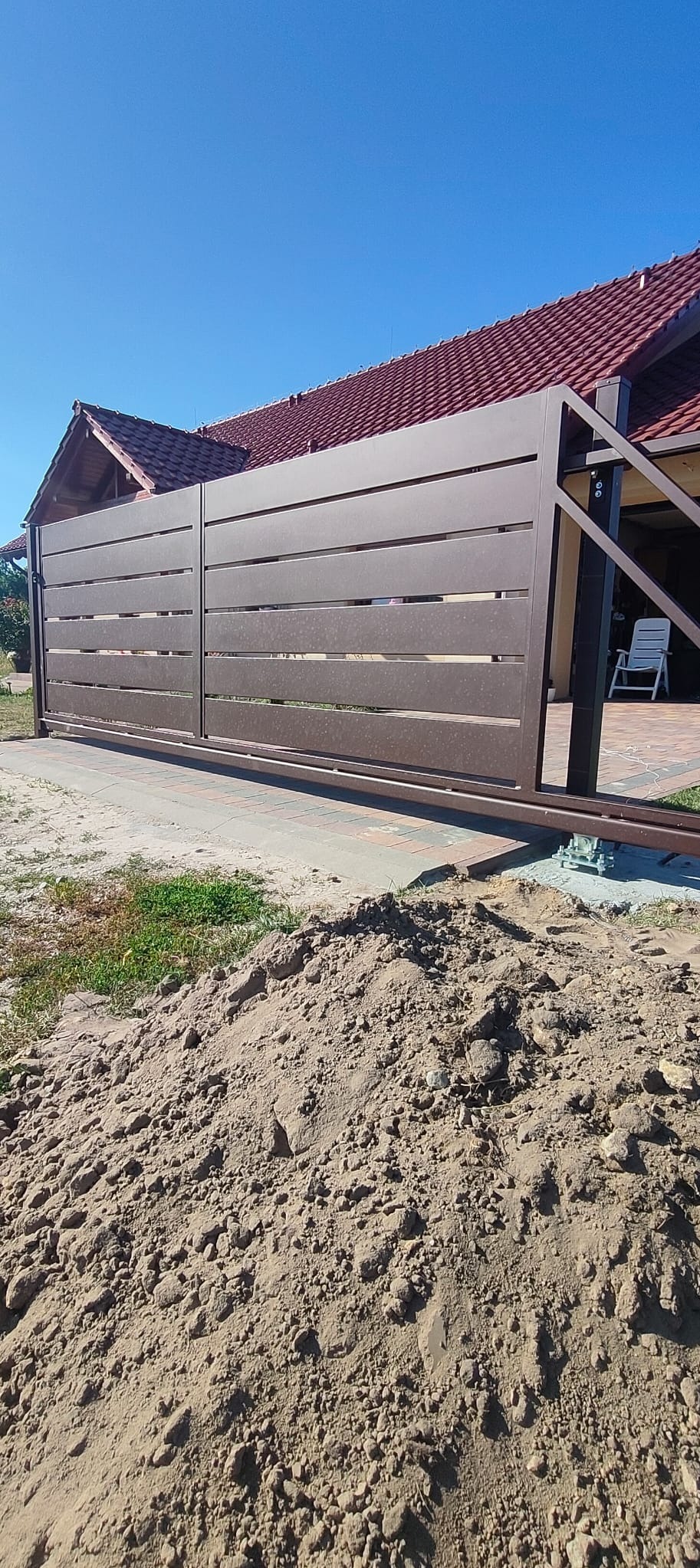 Nowoczesna, przesuwna brama w kolorze brązowym z poziomymi panelami, zamontowana na posesji z widocznym fragmentem domu i stertą ziemi w pierwszym planie.