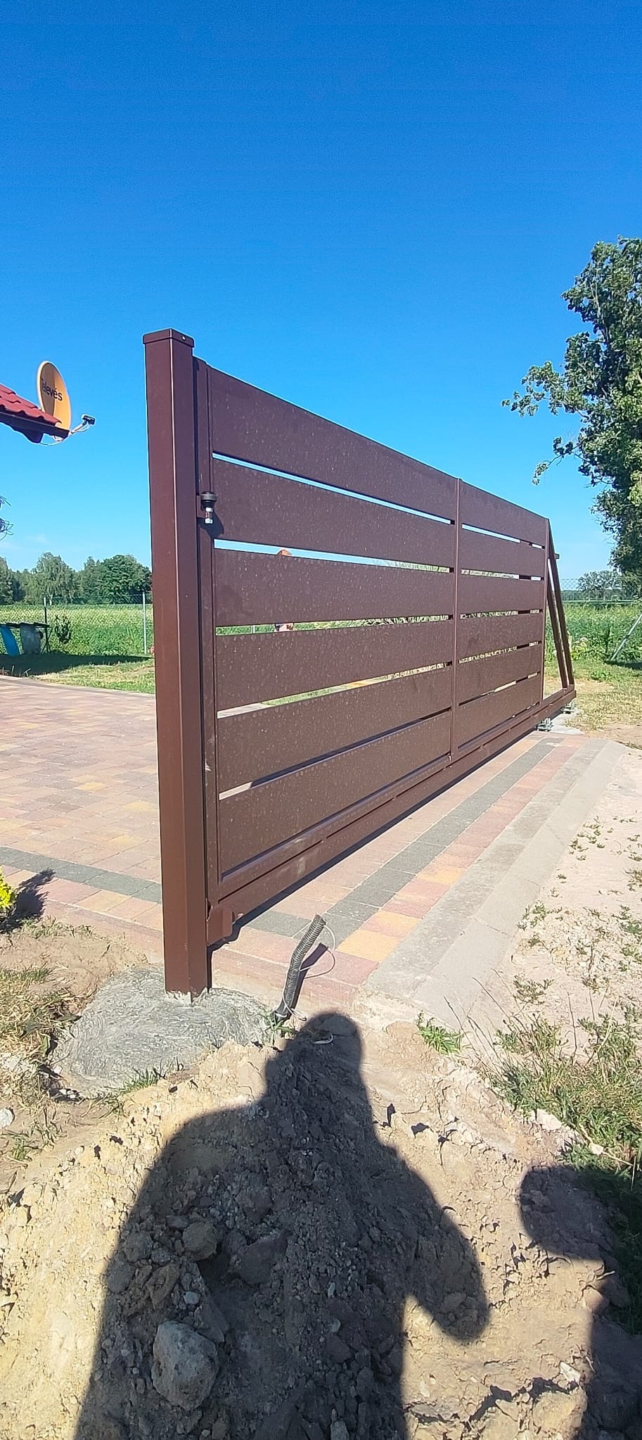 Nowoczesna, przesuwna brama w kolorze brązowym z poziomymi szczeblami, zamontowana na posesji z kostką brukową. Widoczne elementy automatyki i cień osoby.