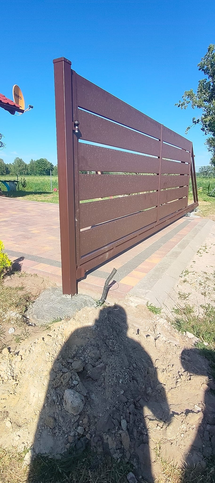 Brązowa, przesuwna brama wjazdowa z poziomymi szczeblami, widoczna w trakcie montażu, z wykopem i kablem w ziemi. Tło: trawnik, drzewa i błękitne niebo.