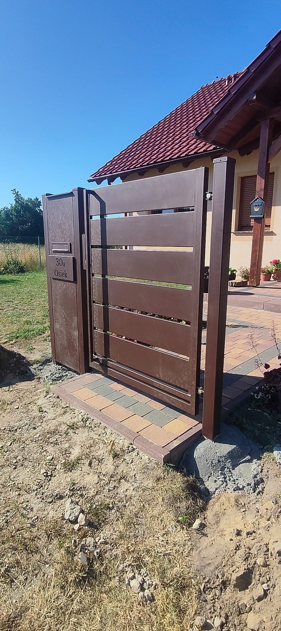 Nowoczesna, brązowa brama posesyjna z numerem domu w Trzebnicy, z poziomymi panelami, zamontowana na kostce brukowej, w słoneczny dzień.