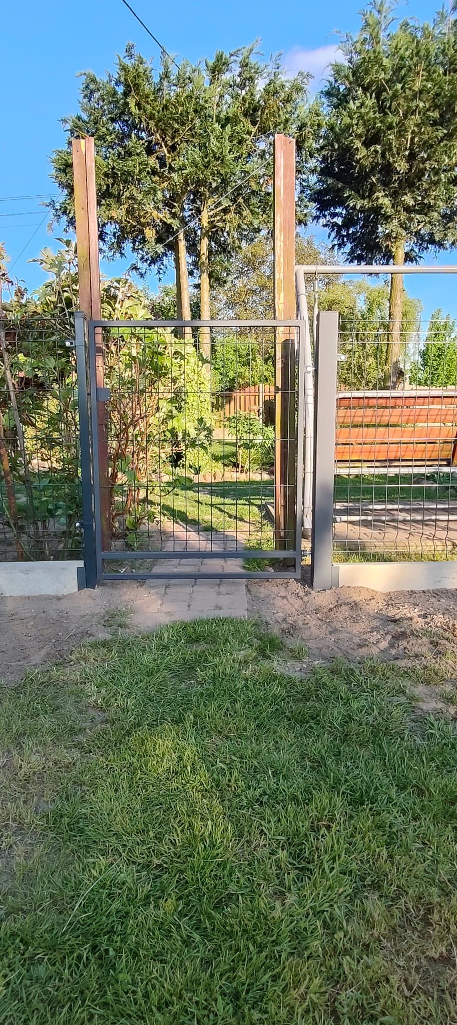 Szara metalowa furtka z siatki na tle zieleni ogrodu, z drewnianymi słupkami i betonową podstawą, widziana z perspektywy trawnika.