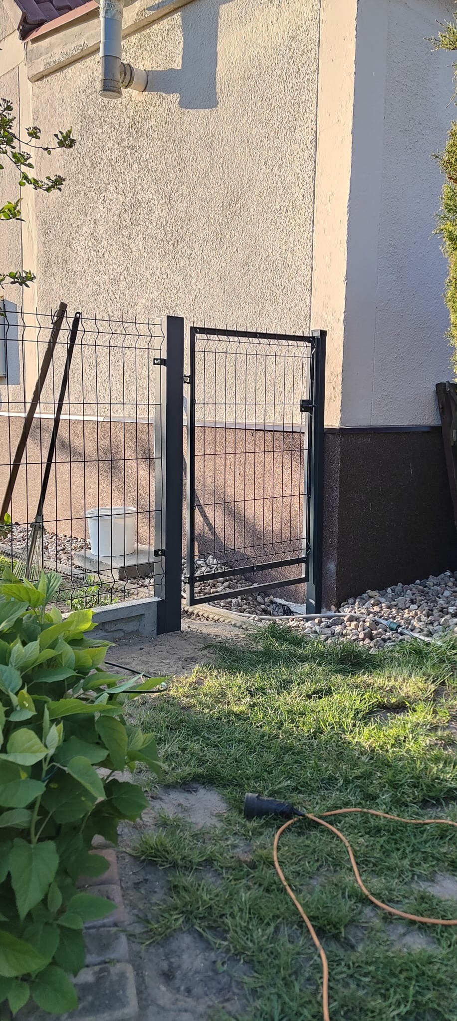 Nowa, czarna furtka z siatki zamontowana przy budynku z elewacją w odcieniach beżu i brązu. Widoczne elementy mocujące i trawnik.