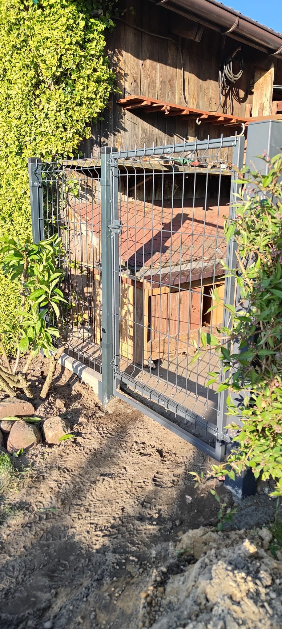Szara siatka ogrodzeniowa z furtką, częściowo zamontowana, widoczna ziemia i roślinność. W tle drewniana budowla z czerwonym dachem.