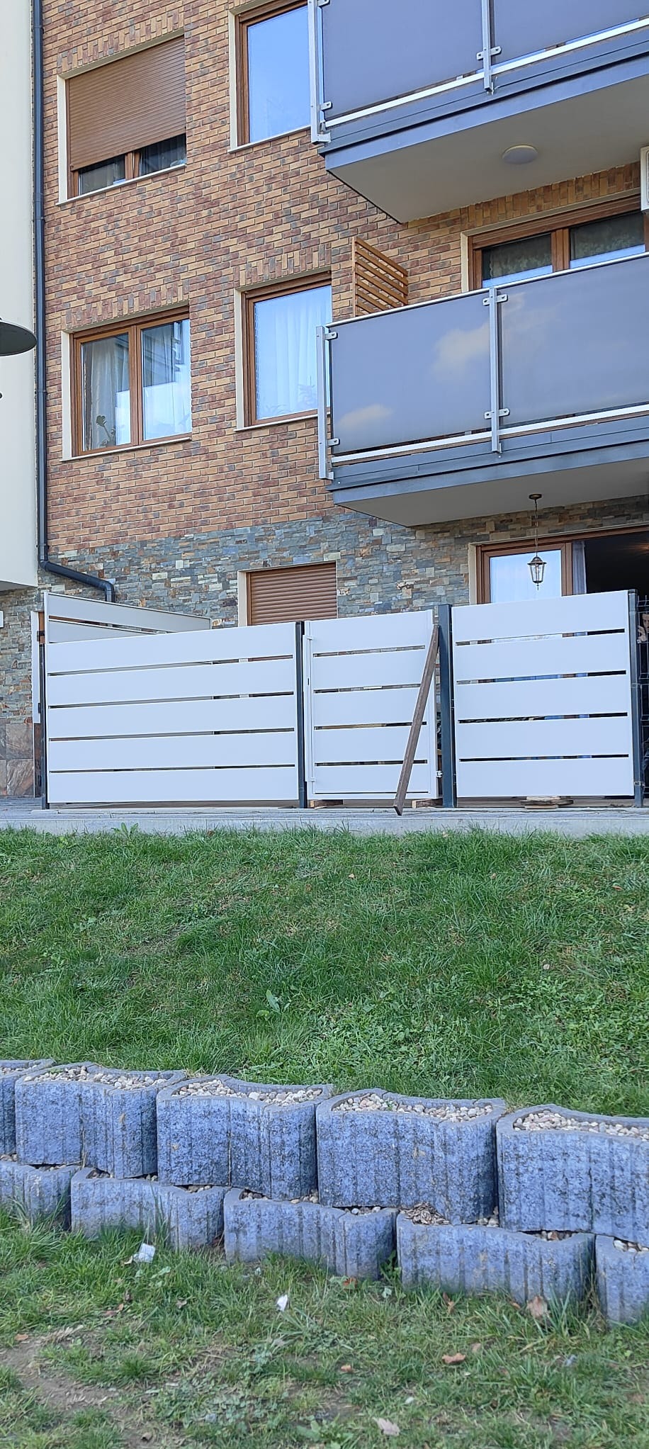 Nowoczesne, białe panele ogrodzeniowe z poziomymi szczeblami, zamontowane przed budynkiem z cegły i kamienia. Widoczne elementy montażowe i trawa.