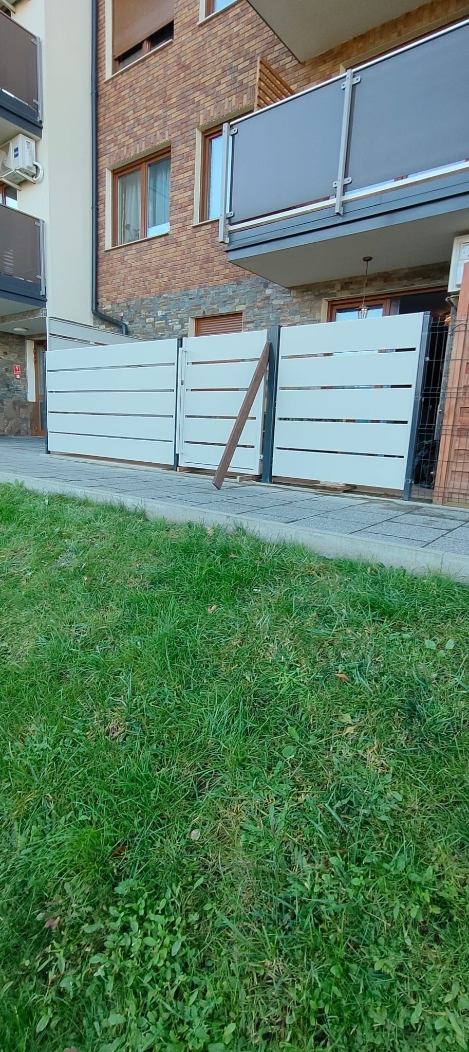 Nowoczesne, poziome przęsła ogrodzeniowe w jasnym odcieniu szarości, z widocznym drewnianym wspornikiem, na tle budynku z elementami kamiennej elewacji.