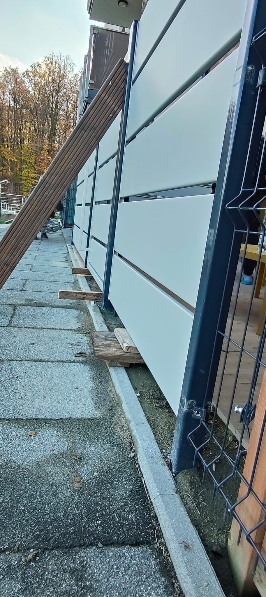 Nowoczesne, białe panele ogrodzeniowe z czarną ramą, w trakcie montażu. Widoczne drewniane podpory i fragment posesji w tle. Ujęcie z dołu, podkreślające perspektywę.