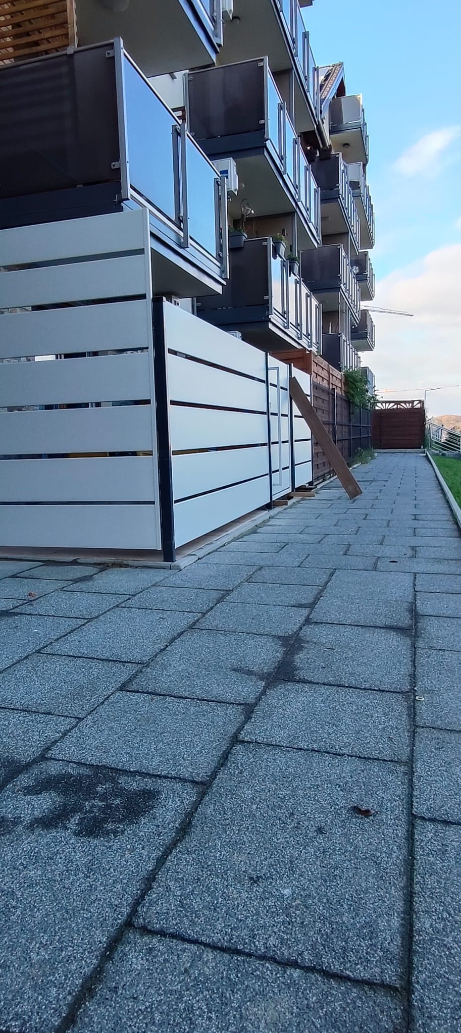 Nowoczesne, poziome panele ogrodzeniowe w kolorze jasnoszarym, zamontowane przy chodniku z kostki brukowej, z balkonami w tle. Widok z dołu, lekko zniekształcona perspektywa.