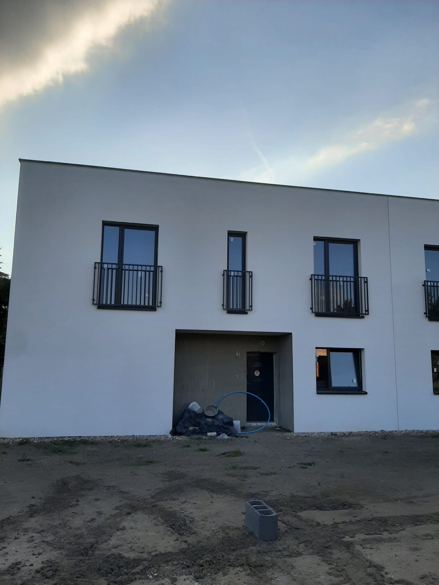 Nowoczesny budynek z balkonami zabezpieczonymi czarnymi, prostymi barierkami. Widoczne materiały budowlane przed wejściem. Minimalistyczny design.