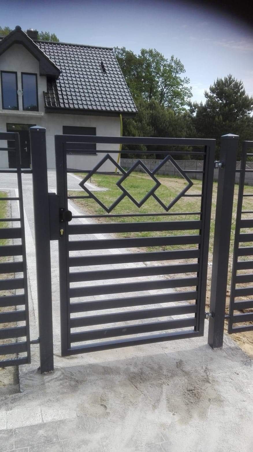 Nowoczesna, grafitowa furtka z poziomymi profilami i dekoracyjnymi rombami, na tle domu z szarą dachówką i zielenią. Minimalistyczny design.