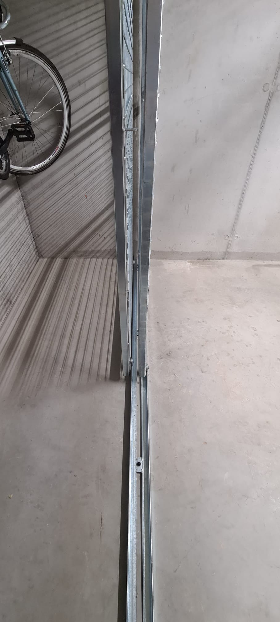 Metalowa konstrukcja z siatką, prawdopodobnie element bramy lub ogrodzenia, widoczna z bliska w surowym wnętrzu. Obok zaparkowany rower.