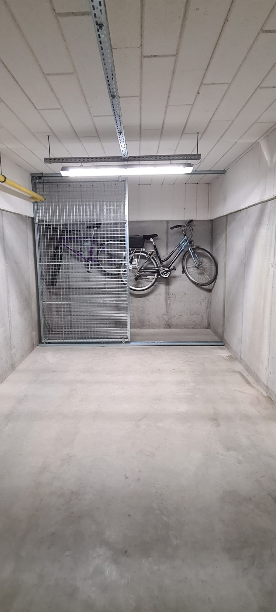 Metalowa, przesuwna krata zabezpieczająca wnękę w betonowej ścianie, wewnątrz rowery. Oświetlenie jarzeniowe. Minimalistyczny, surowy styl.