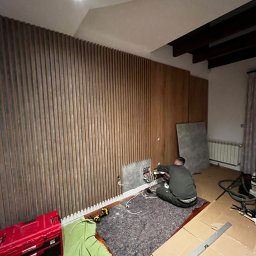 Stolarstwo - Montaż drewnianej ściany dekoracyjnej z pionowych listew. Elektryk pracuje nad instalacją elektryczną. Widoczna skrzynka narzędziowa i poziomica.